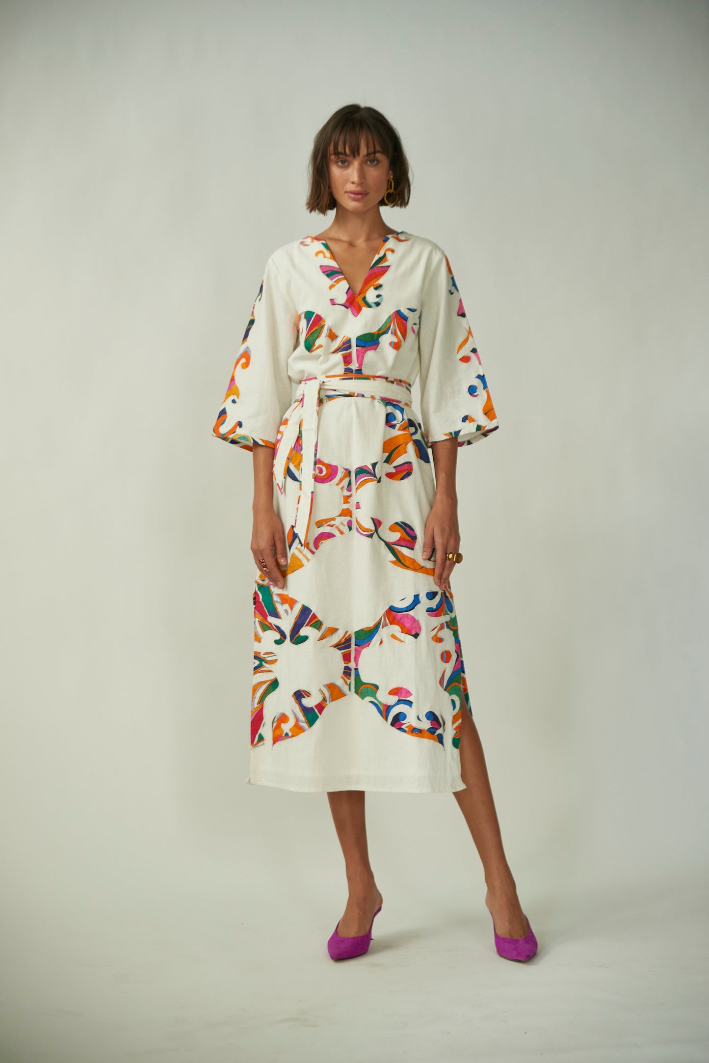 A model wearing a white 'Calliope Kaftan' with a multicolored floral print, V-neckline, three-quarter-length sleeves, cinched waist with a matching fabric belt, and side slit.