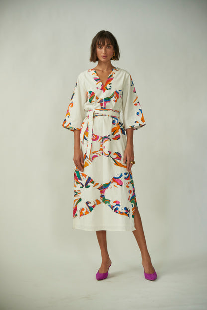 A model wearing a white 'Calliope Kaftan' with a multicolored floral print, V-neckline, three-quarter-length sleeves, cinched waist with a matching fabric belt, and side slit.